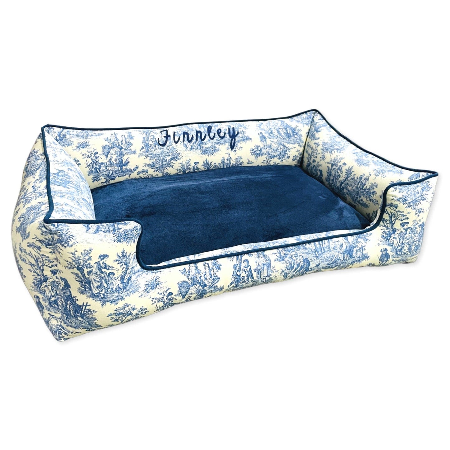 A rectangular bolster dog bed with a toile pattern in blue and white colors, featuring a raised cushioned area and a flat sleeping surface, with a name embroidered on the bolster.