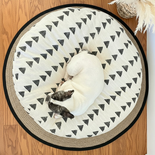 Round boho dog bed with black triangle pattern on a wooden floor, featuring a dog curled 
