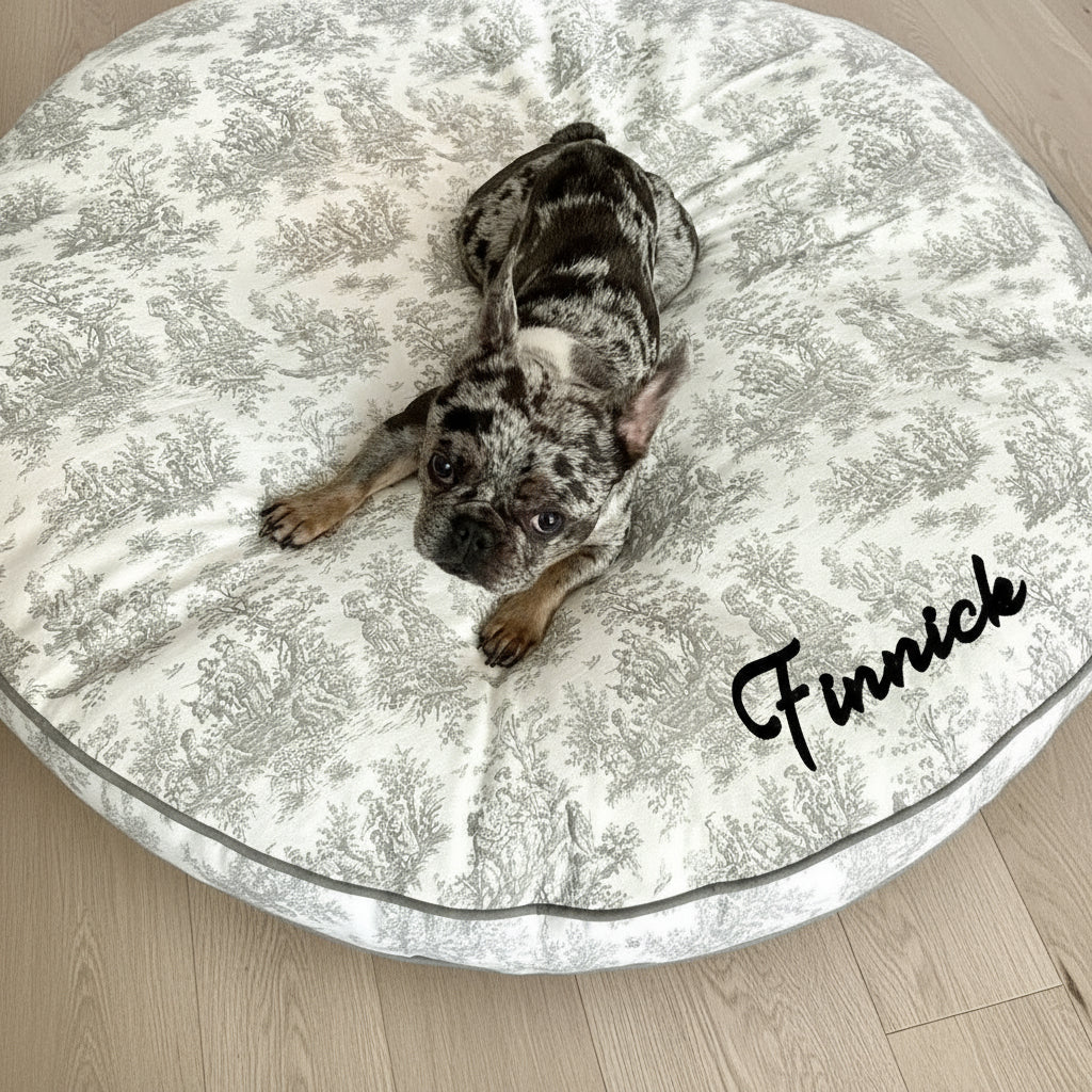 French Bulldog lying on a grey circular dog bed with a toile print and grey trim
